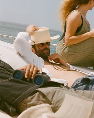 A woman sits to look at the view and a man puts on a white sun hat as a group of friends relaxes on the sundeck of a boat.