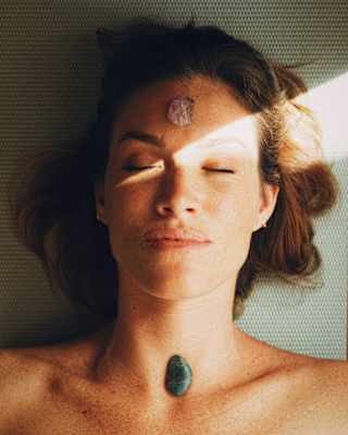 A shaft of light crosses the face of a woman relaxing at the spa with a green stone on her neck and a shell on her forehead.