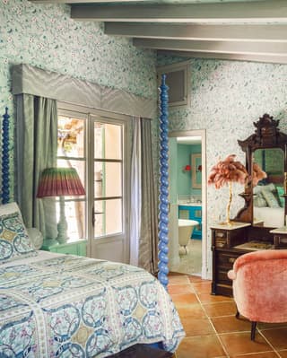 A playful bedroom evoking peacock colours, with blue and green floral wallpaper and bed linens, and green glass lamp stand