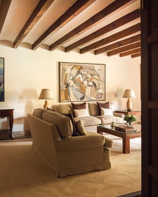 Two cream sofas sit on a large rug in a spacious sitting room with exposed beams, chunky wood tables and abstract art