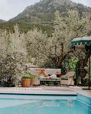 A woman lies on a garden sofa with a sunhat covering her face, seen over a pool with a backdrop of olive trees and mountains.