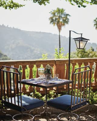 A table and chairs, fashioned from ornate ironwork and soft cushions, is set for 2 beside a stunning view of the mountain