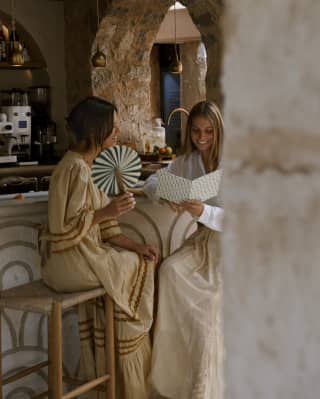 A female guest fans herself as another looks at a menu, seated on stools at Tramuntana Grill, seen through the stone arches.