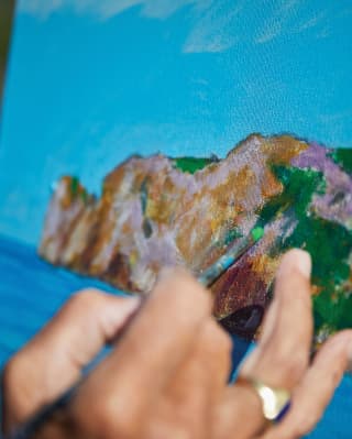 Azure and cobalt meet on the horizon line as a hand with green-tipped paintbrush adds foliage to a canvas's painted hills