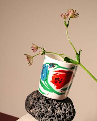 A white mug with colourful abstract designs is balanced on a black lava rock and a white stone, with a sprig of delicate pink flowers placed inside the mug, against a neutral background.
