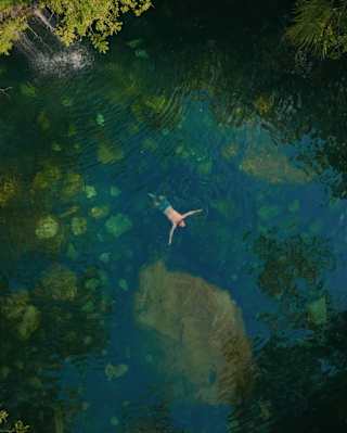 A person floats on their back in a clear, blue-green natural pool surrounded by lush, dense green trees, seen from above. Rocks and underwater features are visible beneath the surface.