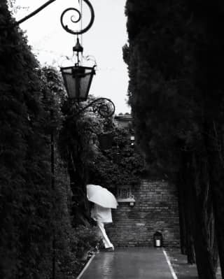 A guest dressed in white, carrying a white umbrella, disappears down a passageway in the hotel grounds, in black and white.