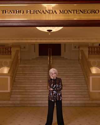 An elderly woman with white hair stands smiling in front of a grand staircase under a sign that reads Teatro Fernanda Montenegro. The elegant setting features ornate railings and warm lighting.