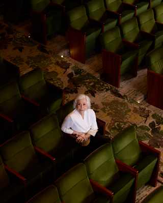 An elderly person with short white hair and a white shirt sits alone in a row of green theatre seats, looking up towards the camera. The surrounding seats are empty.