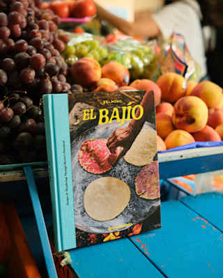 The El Bajío cookbook, with a cover photo of four heated tortillas, rests on a blue market table by an abundant fruit stall.