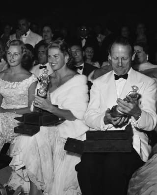 Ingrid Bergman, wearing white fur, and Sir Ashley Clarke hold their awards at the Taormina Film Festival, in black and white.