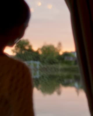 A woman stands at a window, gazing out at the waterway and the sun setting behind it, seen from behind in soft-focus.