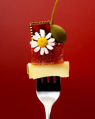Artistic food image of an upright fork, prongs spearing a cube of cheese, topped with a fruit jelly, daisy flower, caper and gem.