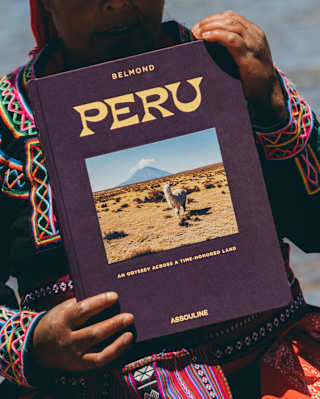 A Peruvian woman in bright traditional woven attire holds a copy of Peru, with a cover-photo of an alpaca, seen close-up.