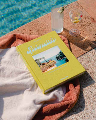 A copy of Splendido, with yellow binding and cover-photo of a bay view rests on a towel with two drinks by a glittering pool.