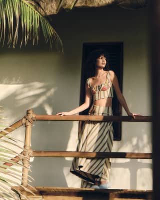 A model in a cream and brown striped skirt and embroidered crop-top by Sandra Weil poses on a raised wooden deck.