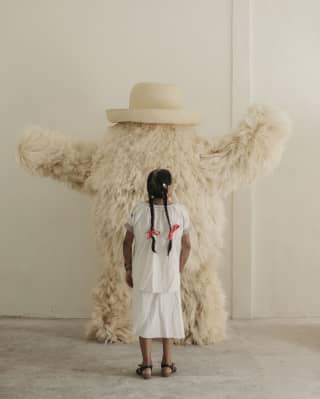 Seen from behind, a girl with long braids looks at a white fluffy monster designed by Fernando Laposse using agave fibres.