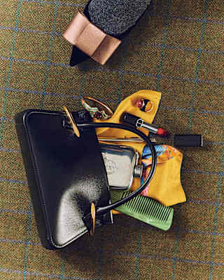 A black handbag is open on a checked surface, revealing a yellow glove, green comb, perfume bottle, sunglasses, and a charging cable. A person wearing grey shoes with a pink bow is partially visible at the top.