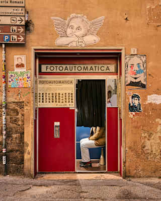 A person sits inside a red photo booth with a black curtain, set in an old, orange wall decorated with graffiti, posters, and a white angel illustration above the booth labelled Fotoautomatica.