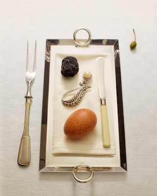 Looking down on a tray with an antique knife, egg, black truffe and vintage brooch at a table with a pickle fork and caper.