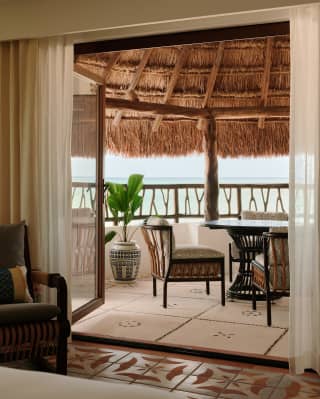 Looking through the doors of an Ocean View Junior Suite to a balcony with a straw canopy, seating and views onto the ocean.
