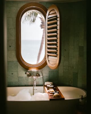 A shutter opens to reveal sea views through an oval window set in the wall above a bubbling bath with a cross-tub soap tray.