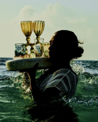 A woman carries a round tray of drinks and unusual glasses on her right shoulder as she wades through waist-high ocean water.