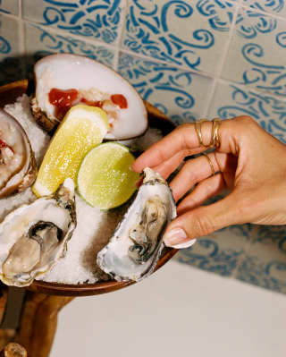 A woman's hand selects a fresh oyster from a selection, presented on ice with lime and lemon on a carved wooden stand.
