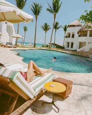 A lone swimmer enjoys the rippling blue tropical pool, seen over a lounger where a female sun-bather's legs stretch out.