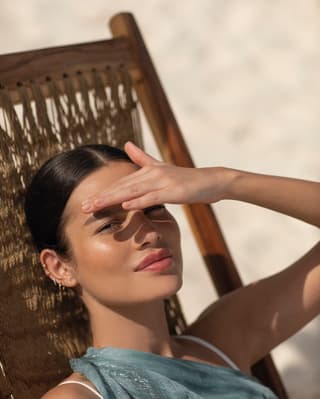 A woman laying on a beach lounger holding up her hand over her face looking at camera directly 