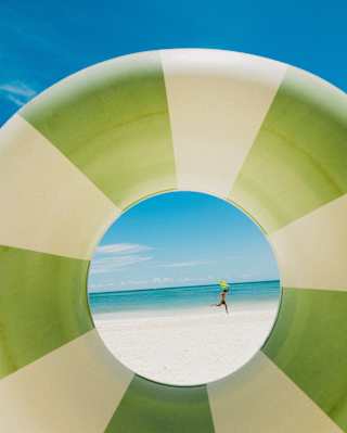 A person is jumping on a sandy beach, seen through the centre of a large green and white striped inflatable swimming ring with the sea and blue sky in the background.