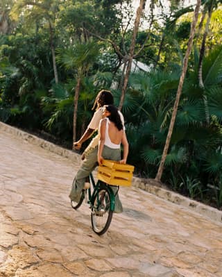 A woman rides on the back of a bike, holding onto a yellow box behind her as the male rider sets off along the stone path.