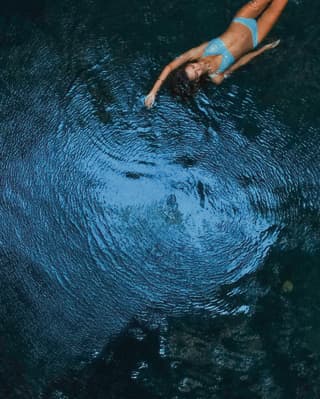 Aerial view of a woman in a blue bikini, swimming on her back with one arm above her head through a sea pool of sunlit water.
