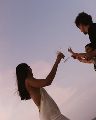 Angled shot of a long-haired woman in a white dress as she raises a glass with two male friends, taken against a lilac sky.