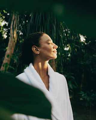 A dark-haired woman wearing a white bath robe closes her eyes as she turns her face to the sun, standing in the garden.