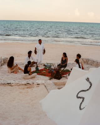 On the beach, a Shaman stands in white, performing a welcoming ceremony for five guests as part of a Temazcal experience.
