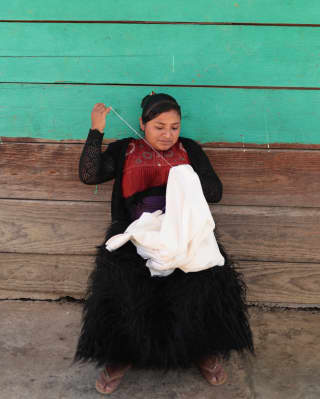 Sitting against a mint and wood plank wall, A local woman stitches with white cotton for fashion brand Collectiva Concepción.