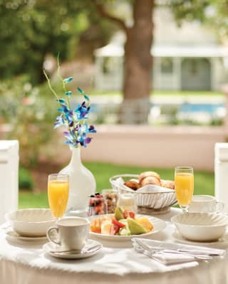 A breakfast table, ready with fruit, juice, pastries and flower vase on the veranda of a Garden Cottage overlooking the pool.