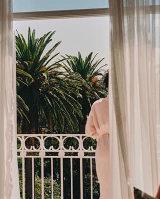 A woman in a pink robe leans on the white railings of her balcony, gazing over lush palm trees, seen from behind a curtain.