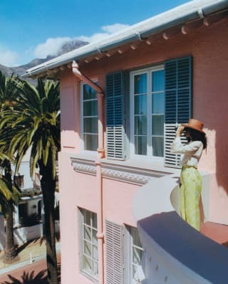 A woman in a brown hat, white shirt and green trousers stands on her suite balcony looking at the view through binoculars.