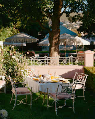 A breakfast table with four chairs, busy with plates and glasses, sits on the lawn in a cottage garden awaiting guests.
