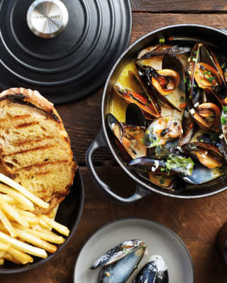 Aerial view of an open mussel pot brimming with shellfish in a buttery liquor with a side bowl of fries and grilled bread.