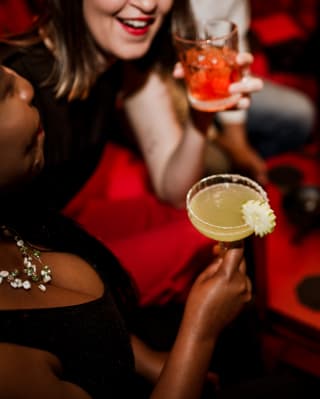 A smiling woman raises her Negroni drink alongside a friend's citrussy cocktail at a busy table in Red Room, seen from above.