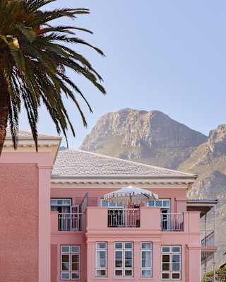 Rocky peaks rise into a blue sky behind a corner of the pink hotel with an upper terrace, part-obscured by a tall palm tree.