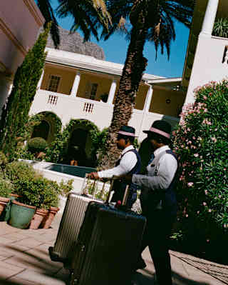 Angled image of two male members of staff, in uniform and hats with pink ribbons, as they wheel suitcases towards the hotel.