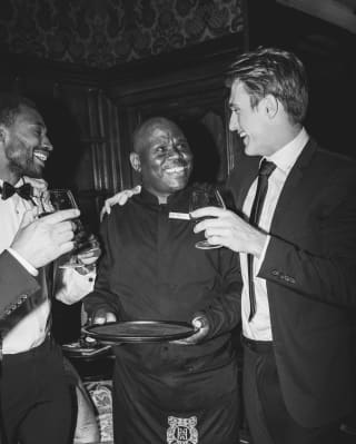 Black and white image of a party as a suited man with a drink rests one arm on the shoulder of a waiter as they share a joke.
