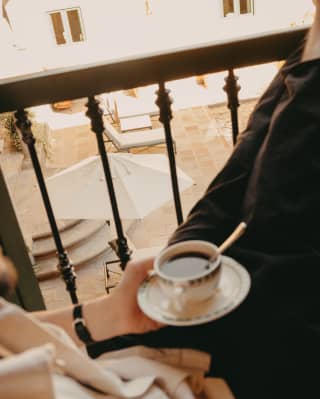 A guest sits with her feet on the balcony rail, holding a cup of black coffee in her lap as she regards the courtyard below.