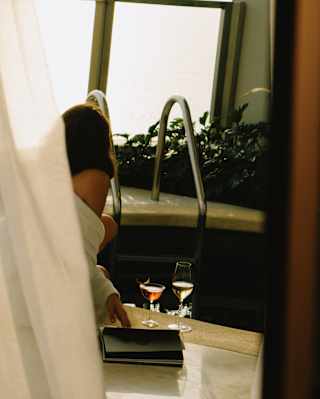 A woman in a robe sits by a private plunge pool where two drinks and a book rest on the edge, part-obscured by a drape.
