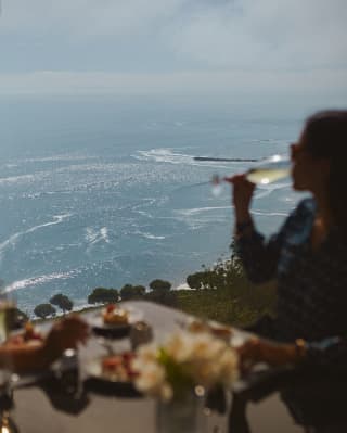 In soft-focus, a woman drinks white wine at an al-fresco table with breathtaking ocean views at the Observatory Restaurant.