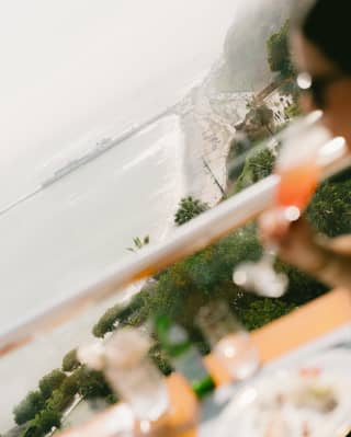 Angled image of a guest sipping a tall cocktail in soft-focus foreground, overlooking the dramatic coast of Lima.